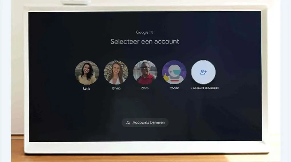 A white-framed screen displays Google TV's 'Selecteer een account' screen with profile icons on a dark background, set on a wooden surface.