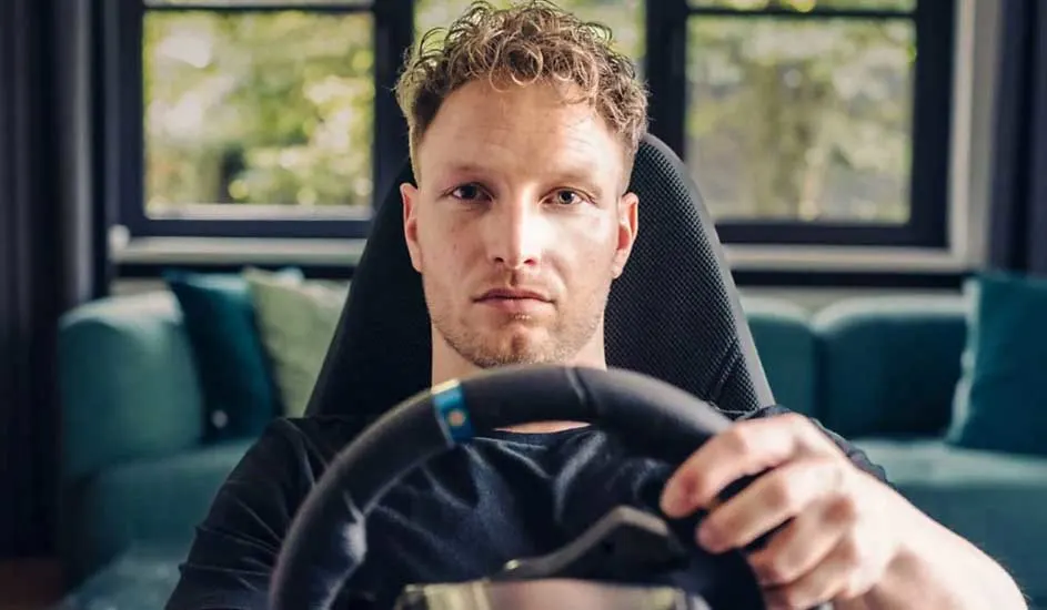 Gros plan sur un volant noir et un siège de course dans un intérieur avec une fenêtre et un canapé bleu-vert en arrière-plan.