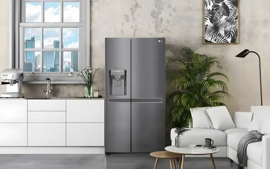 Une cuisine moderne avec coin salon, frigo gris, meubles blancs, grande fenêtre, plantes, canapé et table basse dans une pièce claire.