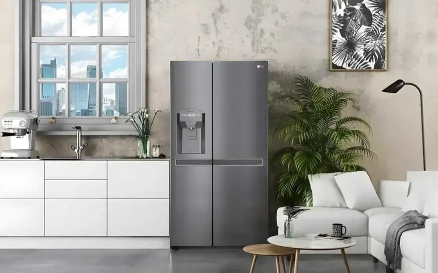 A modern kitchen and sitting area with a grey fridge, white cabinets, large window, plants, sofa and coffee table in a bright room.