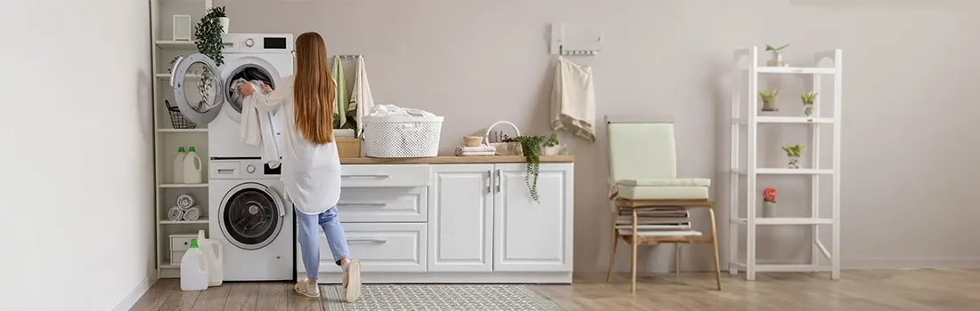 A laundry room with a white washer and dryer, white cabinets, and a shelf. Plants decorate the space.