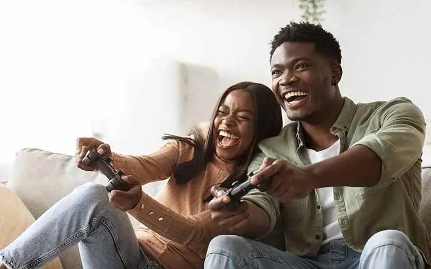 Een man en een vrouw zitten op een grijze sofa, lachend en met gamecontrollers in hun handen.