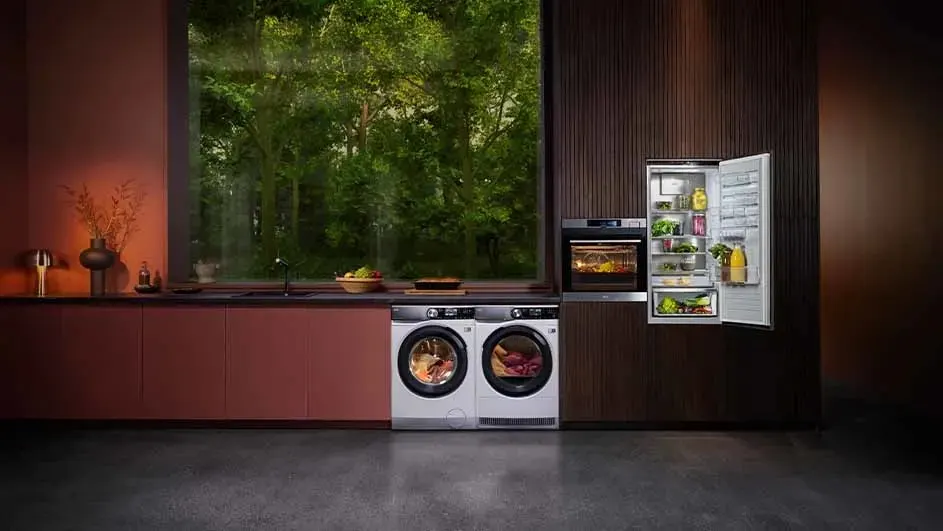 Wasmachines, oven en open koelkast in een moderne keuken met ramen die uitkijken op groene bomen.