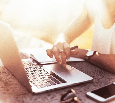 Ordinateur portable argenté, smartphone blanc, lunettes de soleil et carnet blanc sur une table texturée. Lumière vive.