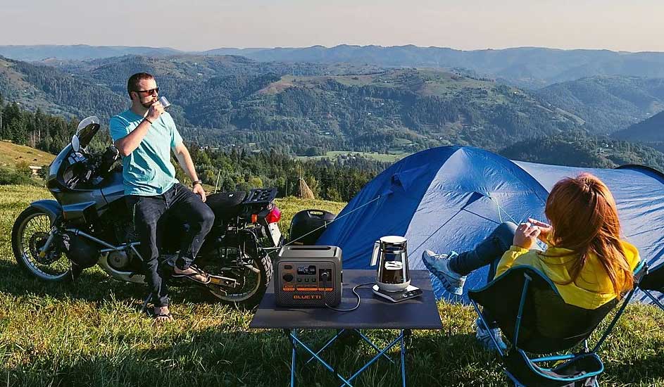 Twee mensen kamperen met een motorfiets, blauwe tent en BLUETTI powerstation op een tafel, omgeven door bergen en groen landschap.