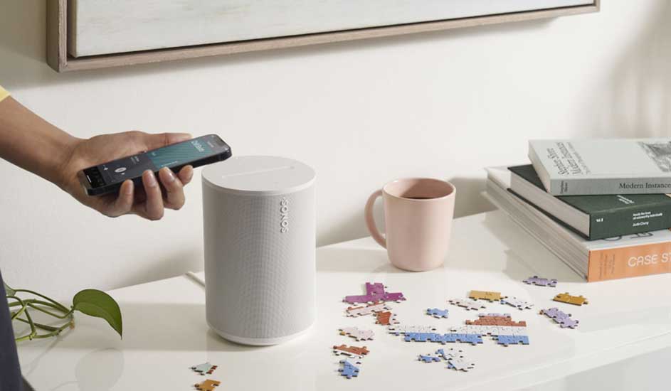 Een tafel met een witte luidspreker, een roze beker, een stapel boeken en verspreide puzzelstukjes. Iemand houdt een telefoon vast.