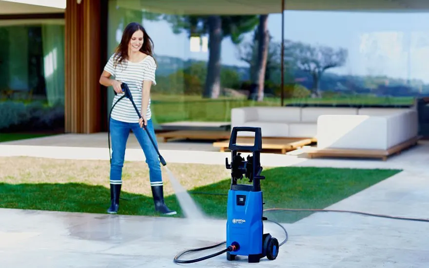 Une femme en chemise rayée utilise un nettoyeur haute pression bleu sur la terrasse d'une maison moderne avec de grandes baies vitrées et montagnes.