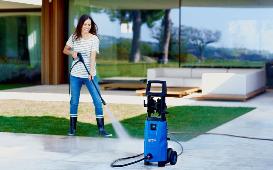 Een vrouw in gestreept shirt gebruikt een blauwe hogedrukreiniger op het terras van een modern huis met grote glazen ramen en bergen op de achtergrond.