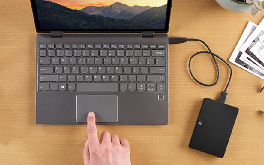 Grijze laptop op een houten tafel met een externe harde schijf aangesloten. Op het scherm is een berglandschap te zien.