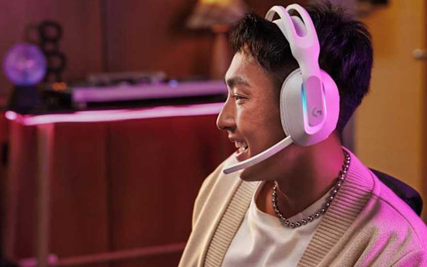Een glimlachende jongeman draagt een wit-roze gaming headset in een sfeervolle kamer met paarse verlichting en gaming-apparatuur.