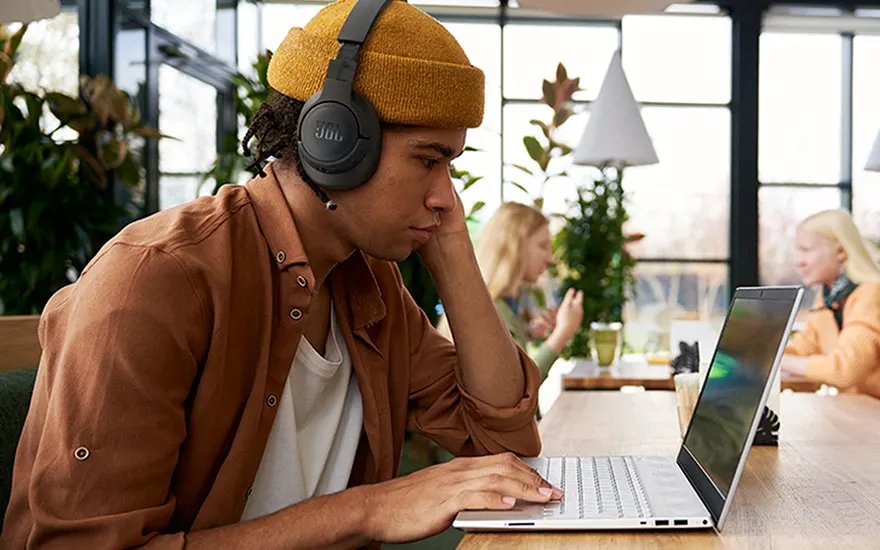 Un homme avec un casque audio travaille sur un ordinateur portable dans un café lumineux. Des plantes et d'autres personnes sont visibles à l'arrière-plan.
