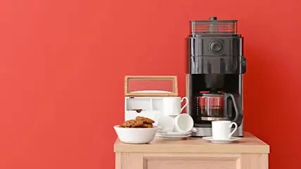 Cafetière noire sur une table en bois avec des tasses, des biscuits et un mur rouge en arrière-plan.