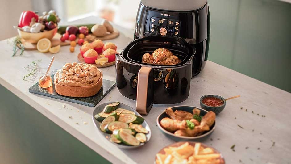 A black Philips air fryer sits on a counter with cooked chicken and fries. There's also a cake, zucchini, and a bowl of mixed vegetables.