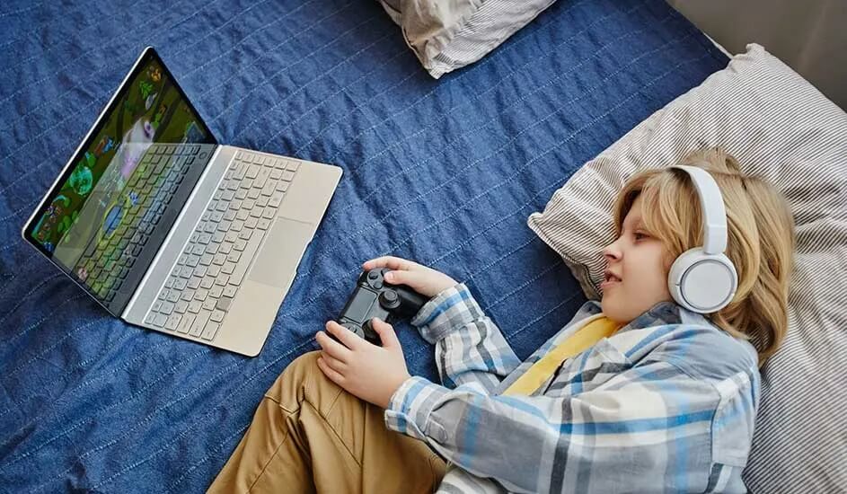 Un enfant allongé sur un lit bleu, casque blanc sur les oreilles, joue à un jeu sur ordinateur avec une manette en main