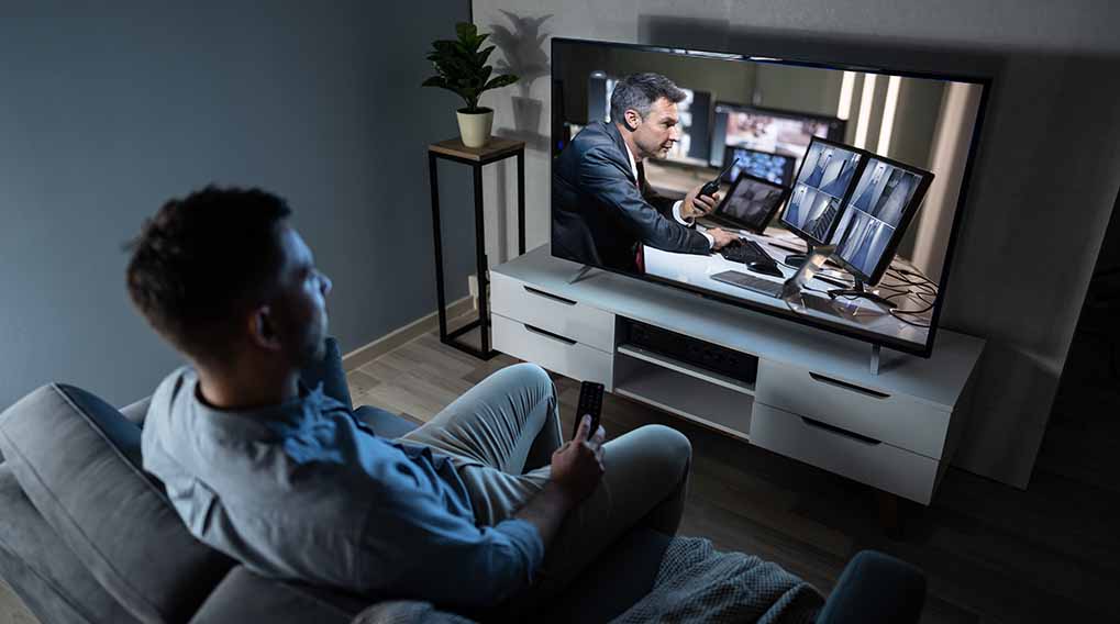 Man op grijze sofa kijkt tv. Op tv: man achter bureau met meerdere monitoren. Naast tv: plant op bijzettafel.