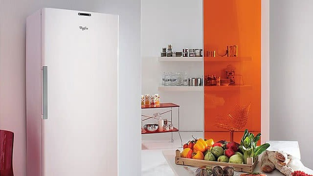 Un frigo blanc près d'étagères avec des aliments et un placard orange. Une caisse de légumes frais est visible au premier plan.