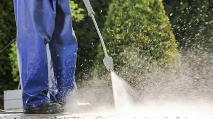 Homme en salopette bleue nettoyant une surface avec un nettoyeur haute pression. Des arbres sont visibles à l'arrière-plan.