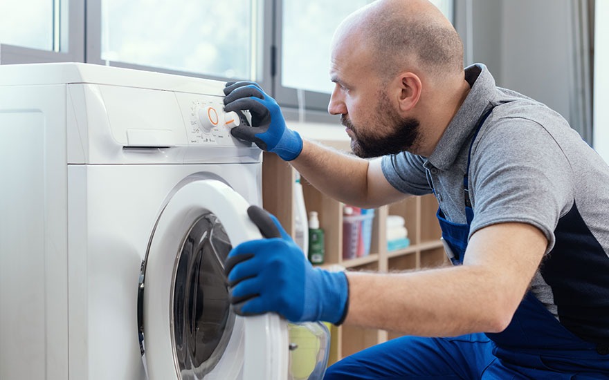 Een witte wasmachine staat tegen een muur bij een raam. Iemand met blauwe handschoenen opent de deur. Op de achtergrond staat een houten rek.
