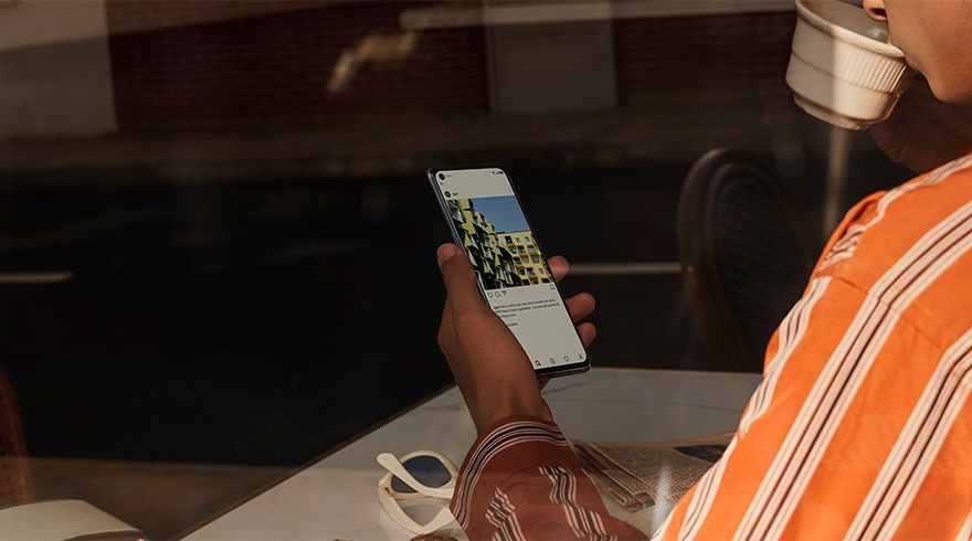 Een hand houdt een smartphone vast met een foto van een gebouw. Op de tafel liggen een zonnebril en een krant.