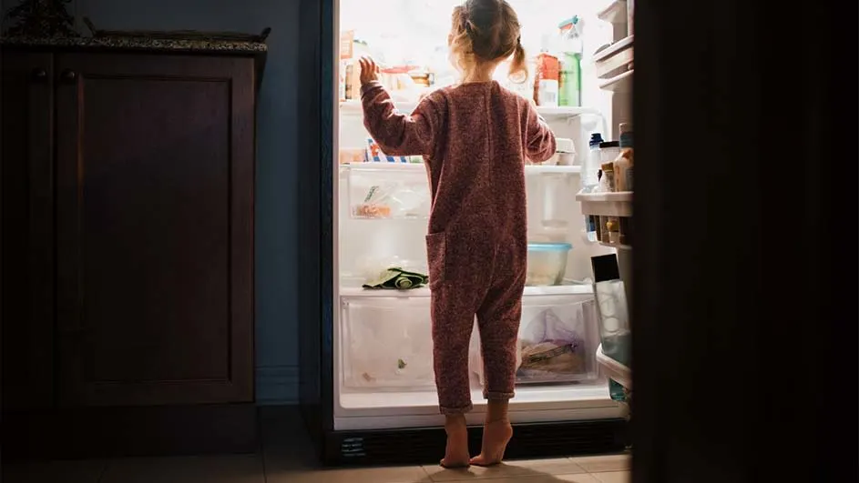 Une personne en pyjama rose devant un frigo ouvert et éclairé, avec des aliments visibles. Une armoire sombre est visible à gauche.
