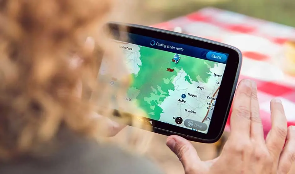 Une personne aux cheveux bouclés tient un GPS affichant une carte et le message « Finding scenic route » pendant un trajet détente en plein air.