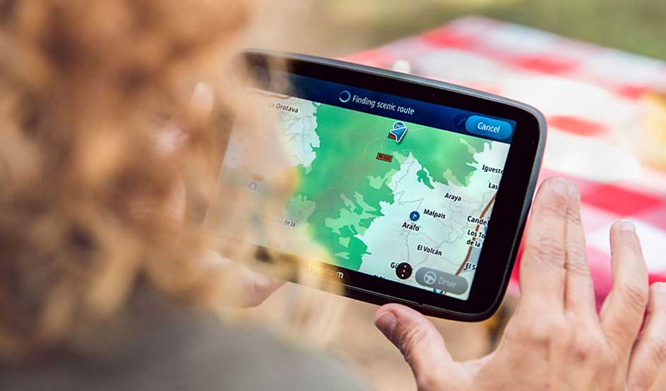 Iemand met krullend haar bedient een GPS-navigatiesysteem met kaartweergave en de melding “Finding scenic route” tijdens een ontspannen trip.