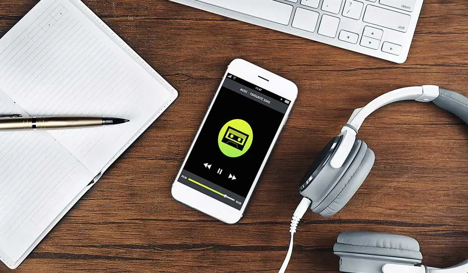 Een bureau met een notitieboek, pen, telefoon met muziek-app, witte toetsenbord en een grijze hoofdtelefoon.