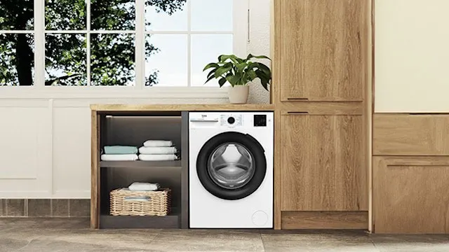 White Beko washing machine in a tidy laundry room with wooden cabinets, folded towels, and a green plant by the window.