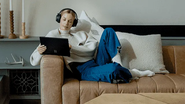 Une tablette noire est tenue sur un canapé en cuir marron avec des coussins blancs. Un casque audio noir est sur le canapé.