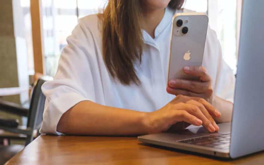 Op een houten tafel staat een grijze laptop en een zilverkleurige smartphone met drie cameralenzen.