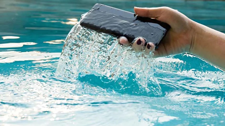 Een hand haalt een zwarte smartphone uit het water van een zwembad, merk is niet zichtbaar.