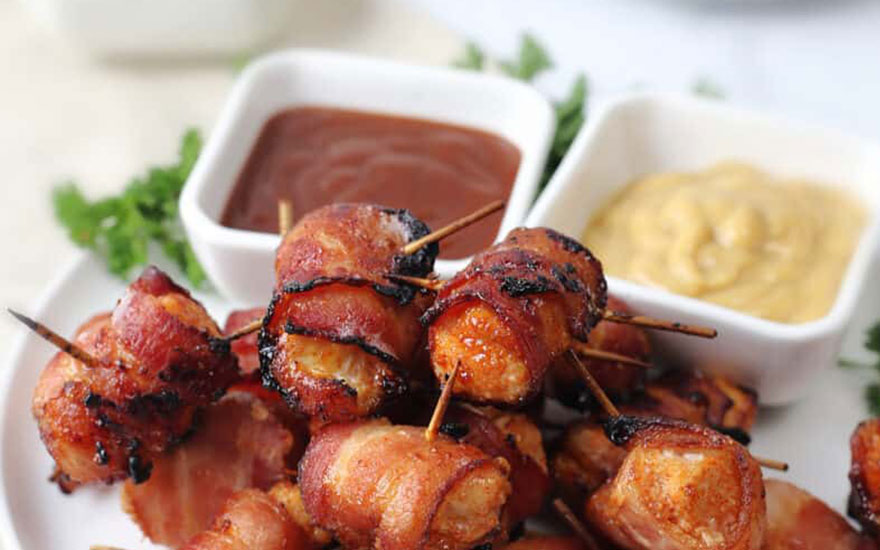 Gros plan d'une assiette de bouchées de poulet enroulées de bacon, fixées avec des cure-dents, avec des sauces à tremper.