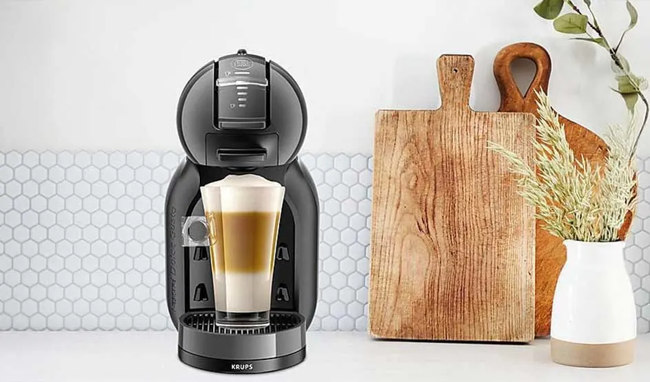 A black Krups coffee machine dispenses layered coffee into a glass. Wooden cutting boards and a vase with wheat decorate the white countertop.