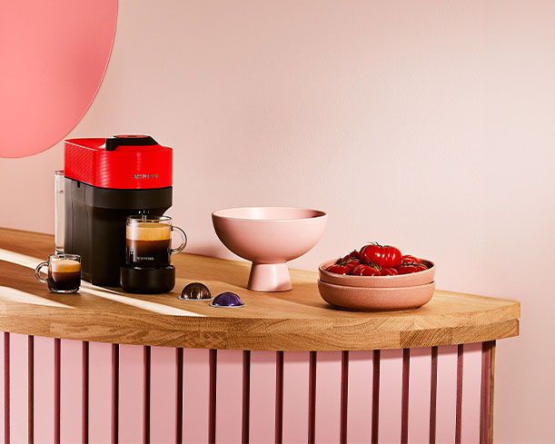 Machine à café rouge et noire, tasses, bols roses et tomates sur comptoir en bois avec base rayée rose.
