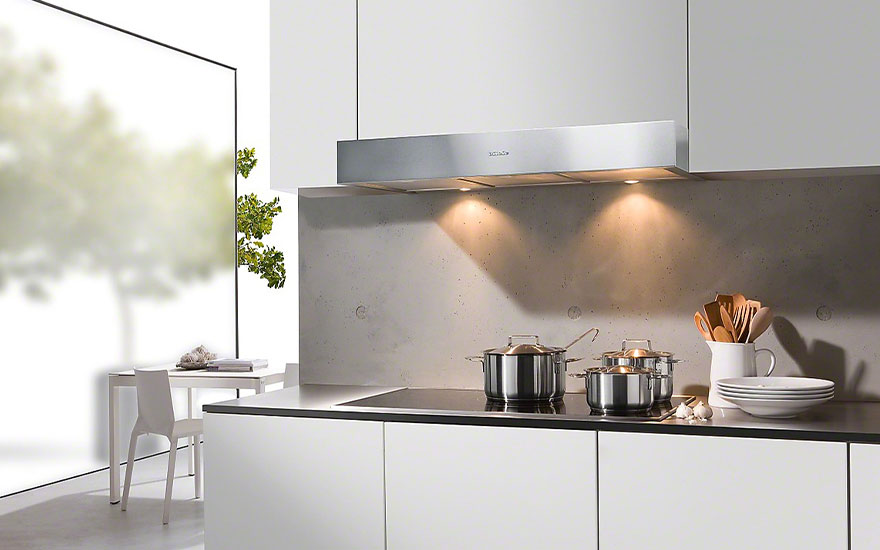 Cuisine moderne blanche avec hotte argentée, casseroles sur plaque de cuisson, ustensiles dans un pot et table près de la fenêtre.