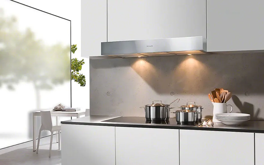 Cuisine moderne blanche avec hotte argentée, casseroles sur plaque de cuisson, ustensiles dans un pot et table près de la fenêtre.