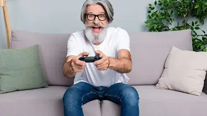 Een man zit op een grijze bank met kussens en speelt een spel met een zwarte controller.