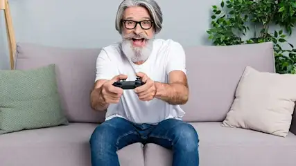 Un homme assis sur un canapé gris tient une manette de jeu noire. Un coussin vert est à gauche et un coussin beige à droite.