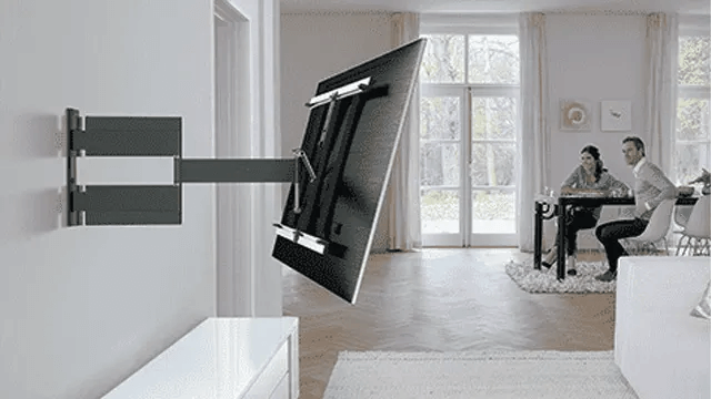 Een grijze tv aan een beweegbare muurbeugel in een kamer met wit meubilair en een groot raam.