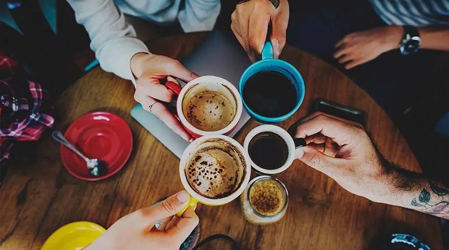 Une table en bois présente des tasses à café colorées, des soucoupes rouge et jaune, et un pot en verre. Des mains tiennent les tasses.