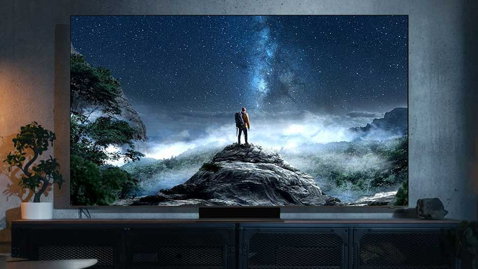 Un grand écran affiche un paysage montagneux nocturne avec une voie lactée brillante, posé sur un meuble bas dans une pièce.