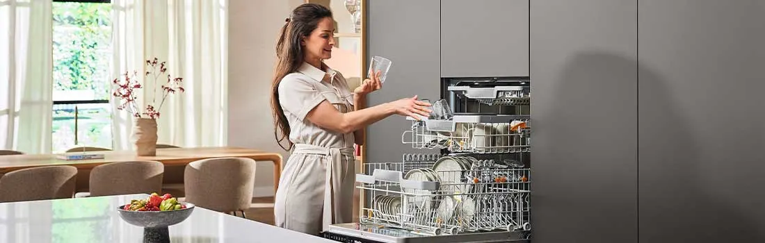 A modern kitchen features a dishwasher filled with dishes and glassware, next to gray cabinets. A fruit bowl sits on a white counter.