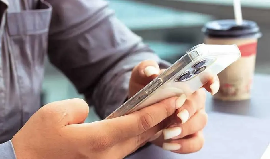 Hands hold a clear-cased Apple iPhone with three rear cameras; long manicured nails visible, takeaway coffee cup blurred in the background.