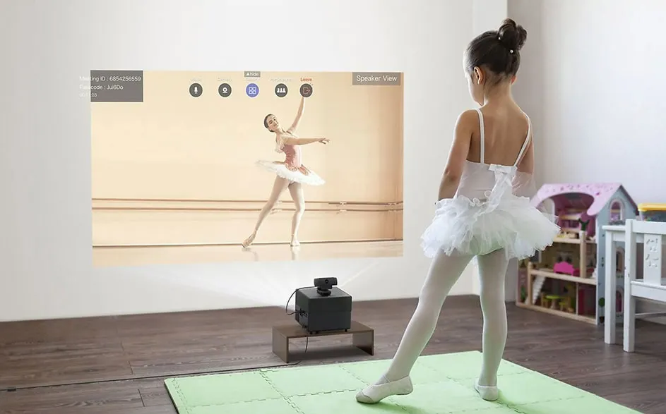 Een projector toont een balletdanseres op een witte muur, met een zwart apparaat op een houten plank eronder. Een groene mat ligt op de houten vloer.
