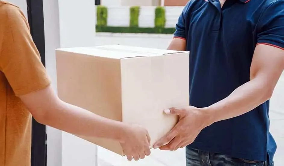 Een beige kartonnen doos wordt door twee paar handen overgedragen. De achtergrond toont een vaag huis met struiken.