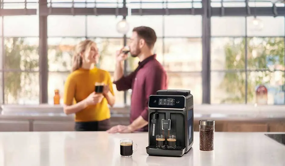 Une machine à café noire sur un comptoir blanc, avec des tasses de café. Un bocal de grains de café est à côté. Grande fenêtre en arrière-plan.