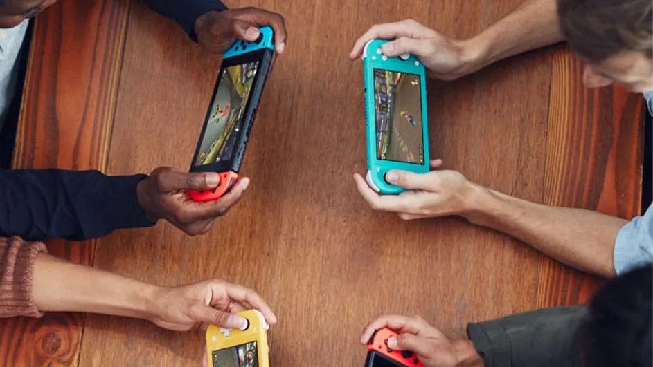 Quatre consoles portables, aux couleurs variées, affichent des jeux. Elles sont tenues au-dessus d'une table en bois marron.