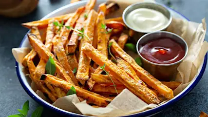 Een blauwe schaal met zoete aardappelfrietjes, ketchup, mayonaise en kruiden op een grijze ondergrond.