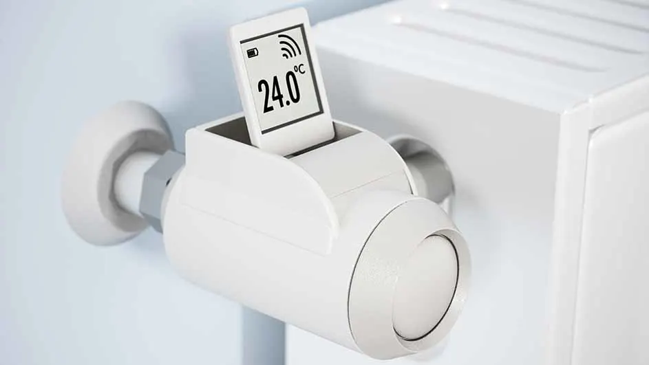 Un thermostat blanc affiche 24.0°C sur un écran. Il est fixé à un radiateur blanc sur un mur clair.