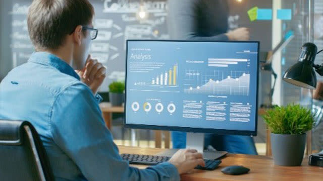 Un écran affiche des graphiques en bleu, avec des histogrammes et des courbes. Un clavier et une souris sont visibles sur le bureau en bois.